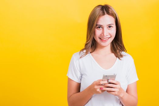 Asian Thai portrait happy beautiful cute young woman smile standing playing game or writing SMS message on smartphone looking to the phone, studio shot isolated on yellow background with copy space