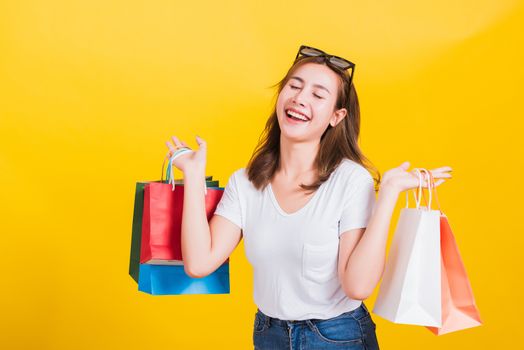 Asian Thai portrait happy beautiful cute young woman smiling stand with sunglasses excited holding shopping bags multi color close eyes, studio shot isolated yellow background with copy space