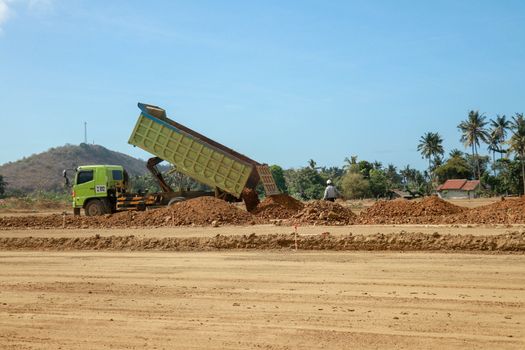 Green dump truck dumps its load of clay on construction site for road construction. Moto GP Mandalika racing circuit under construction, Lombok, Indonesia. Trucking industry, freight cargo transport.