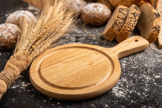 An empty wooden cutting board and wheat on the table with various kinds of bread.