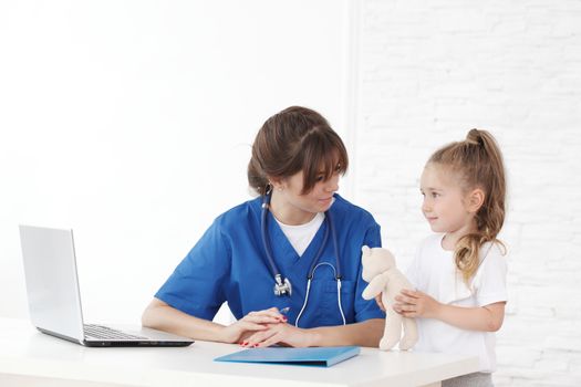 Young smiling female pediatrician doctor and her little patient with teddy bear at clinical office