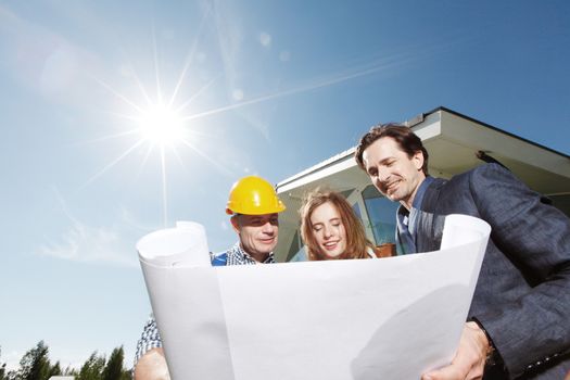 Worker shows construction plan to young couple outside house under construction sun in shining