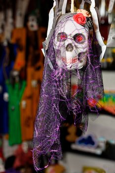 Halloween skull doll with a smile, decorated with feminine makeup, flowers in the form of a hat and a lilac veil, isolated on a blurred dark background.