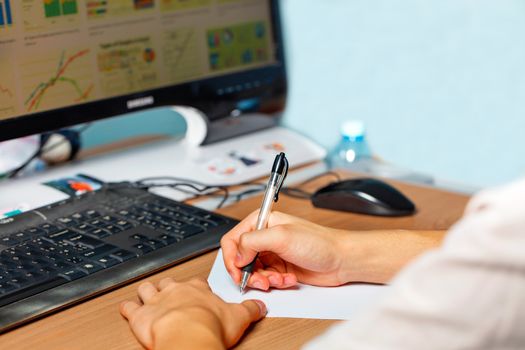 The student's hands hold a pen in front of a blank sheet of paper, sitting at a table in front of a computer monitor, preparing to take notes at a lecture. E-learning, self-education in quarantine. Selective focus, copy space.