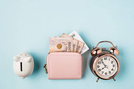 Saving piggy bank with wallet of Thai currency, 1000 Baht, money banknote of Thailand and bell alarm clock on blue background for business, finance and time management concept