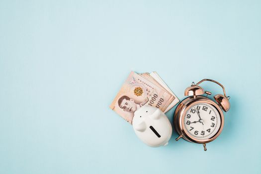 Saving piggy bank with Thai currency, 1000 Baht, money banknote of Thailand and bell alarm clock on blue background for business, finance and time management concept