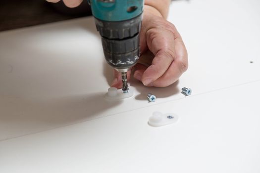 Electric screwdriver with cruciform bat wraps into furniture cabinet, screws lie nearby during assembly of purchased furniture.