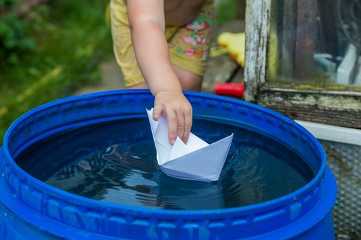 A boy plays with paper boat in the water barrel in the garden. 