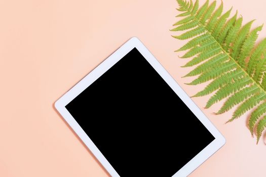 Tablet mockup on beige work desk surrounded with green leaf of fern. Free space beside for text. Flat lay composition.