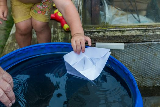A boy plays with paper boat in the water barrel in the garden.