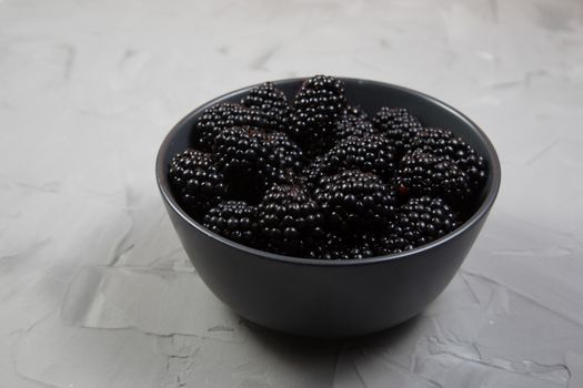 Ripe sweet blackberry in a gray ceramic bowl on gray concrete background.