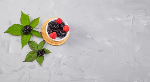 ripe blackberry with leaves on a concrete background, in a white ceramic bowl, top view , place for text, banner