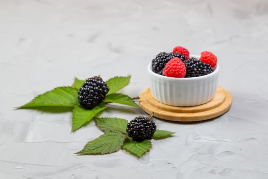.ripe blackberry with leaves on a wooden cutting board in a white ceramic plate on concrete background, selective focus