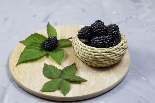 ripe blackberries with leaves in a glass bowl on a bamboo cutting board on a concrete background, rustic, place for text, selective focus