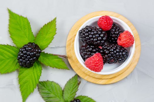 ripe blackberry with leaves on a wooden cutting board in a white ceramic plate on concrete background