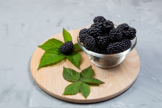 .ripe blackberries with leaves in a glass bowl on a bamboo cutting board on a concrete background, rustic, place for text