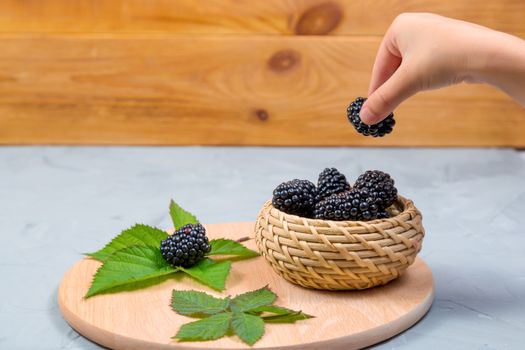 Child's hand takes a blackberry from a wicker bowl on a gray concrete background