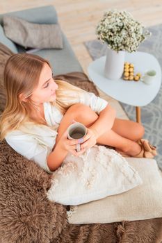 Young beautiful girl sitting on a couch wrapped in cozy blanket with cup of coffee