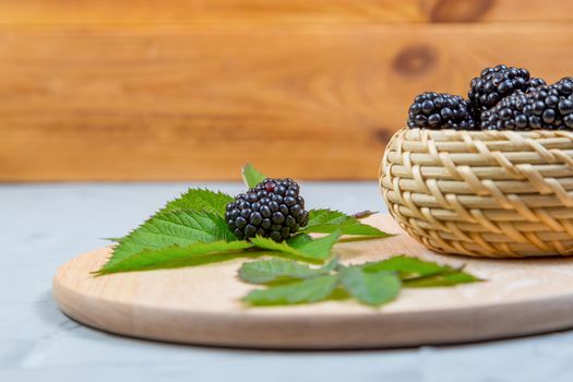ripe blackberry with leaves on a wooden cutting board in a wicker bowl on concrete background.