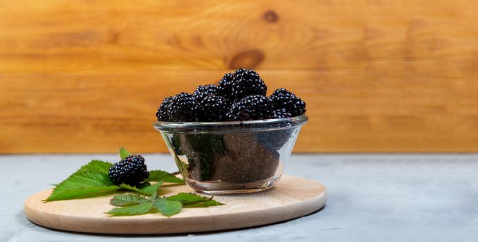 ripe blackberries with leaves in a glass bowl on a bamboo cutting board on a concrete background, rustic, place for text