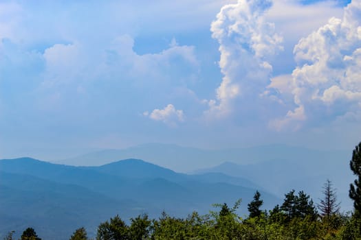 Forest at the bottom. Blue silhouette of a mountain in the distance, with clouds in the blue sky. Mountains and hills in the distance. Bosnia and Herzegovina.