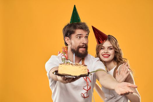 Happy man and woman in a cap celebrating a birthday on a yellow background with a cake in their hands. High quality photo