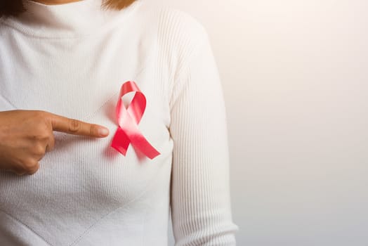 Breast cancer awareness healthcare and medicine concept. Close up Asian woman wear white shirt pointing finger to pink breast cancer awareness ribbon, studio shot isolated on white background