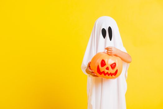 Funny Halloween Kid Concept, little cute child with white dressed costume halloween ghost scary he holding orange pumpkin ghost on hand, studio shot yellow on white background