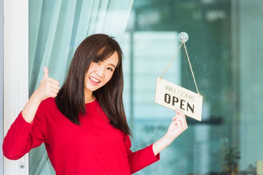 Asian young woman smile show finger thumb up for good sign she notice sign wood board label "WELCOME OPEN" hang through glass door front shop, Business turning open after coronavirus pandemic disease