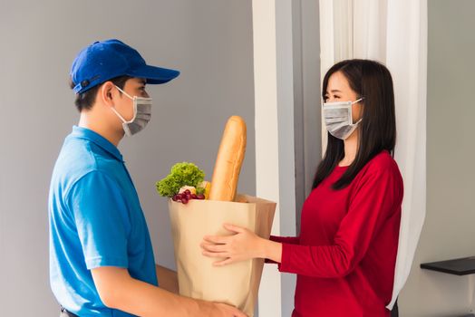 Asian young delivery man in uniform wear protective face mask he making grocery service giving fresh food to woman customer receiving front house under pandemic coronavirus, Back to new normal concept