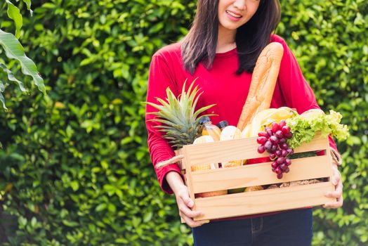 Portrait of Asian beautiful young woman farmer standing she smile and holding full fresh food raw vegetables fruit in a wood box in her hands on green leaves background