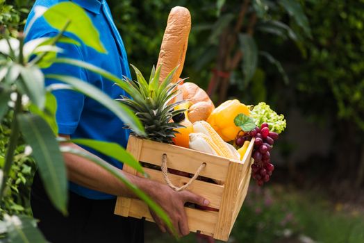 Asian man farmer wears delivery uniform he holding full fresh vegetables and fruits in crate wood box in hands ready give to customer harvest organic food on the garden place green leaves background