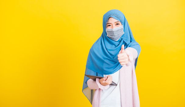 Asian Muslim Arab, Portrait of happy beautiful young woman Islam religious wear veil hijab and face mask protect she quarantines disease coronavirus show finger thumb up for good sign isolated yellow