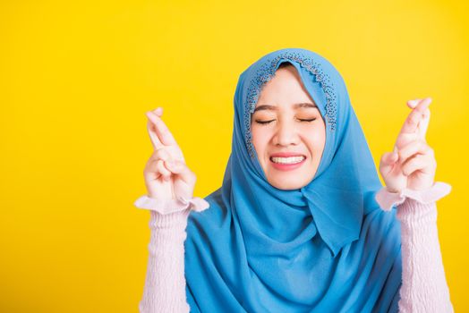 Asian Muslim Arab, Portrait of happy Asian beautiful young woman Islamic religious wear veil hijab she smiling and holding fingers crossed for good luck, studio shot isolated on yellow background