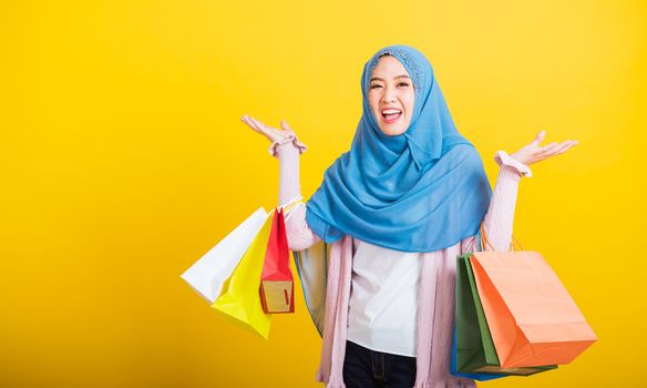 Asian Muslim Arab, Portrait of happy beautiful young woman Islam religious wear veil hijab funny smile she holding colorful shopping bags so glad shopping bags hand raise in studio isolated on yellow