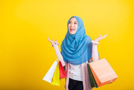 Asian Muslim Arab, Portrait of happy beautiful young woman Islam religious wear veil hijab funny smile she holding colorful shopping bags so glad shopping bags hand raise in studio isolated on yellow