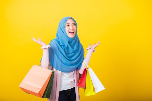 Asian Muslim Arab, Portrait of happy beautiful young woman Islam religious wear veil hijab funny smile she holding colorful shopping bags so glad shopping bags hand raise in studio isolated on yellow
