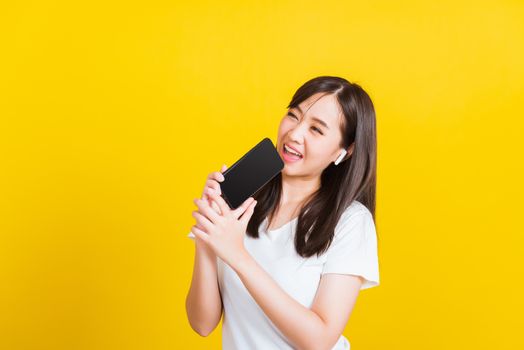 Portrait Asian of a happy beautiful young woman holding a mobile smart phone and wearing wireless headphones listening to music from smartphone studio shot isolated on yellow background