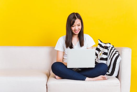 Portrait Asian of happy beautiful young woman work from home she sitting on sofa using laptop computer in house living room studio shot isolated on yellow background