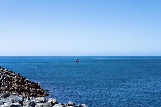 A fishing boat heading out to sea from the harbour rock wall, with a coal tanker on the horizon and another travelling fishing boat