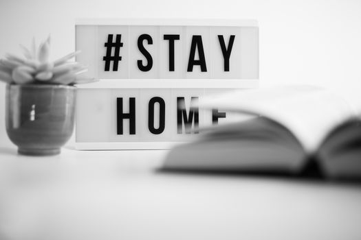 Sign Stay Home, home plant and open book in soft focus on white background. Black and white. Concept of learning, studying at home during coronavirus. Open university, college course, online distance learning