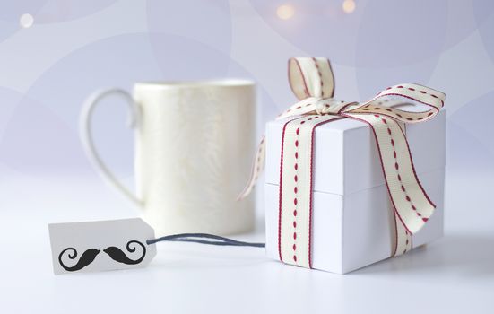 Cup of morning coffee, wrapped gift box, gift tag with icon of moustache on light background with bokeh. Fathers day, birthday concept