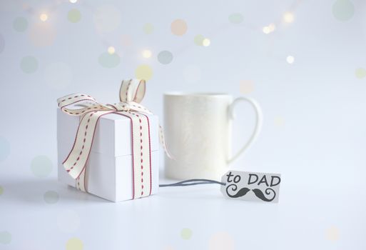 Cup of morning coffee, wrapped gift box, gift tag with word DAD and icon of moustache on light background with bokeh