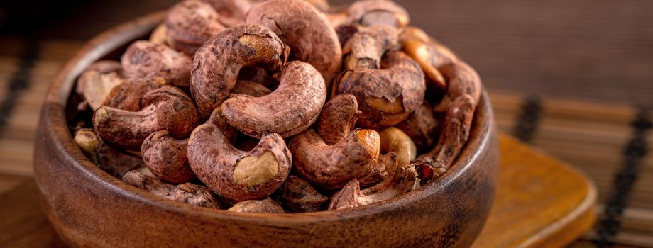 Cashew nuts with peel in a wooden bowl on wooden tray and table background, healthy raw food plate.