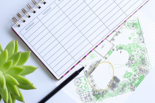 Weekly planner with pen, home plant in soft focus and computer garden design drawing sketch. Landscaping architectural project execution, garden design landscape concept