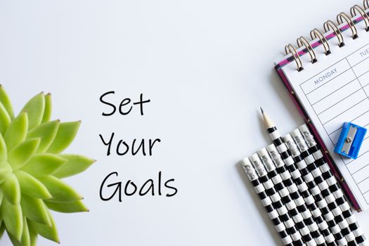 Sign Set Your Goals, home plant soft focus, weekly organiser planner, sharpener and pencils. Back to school. White background. Stationery flat lay.
