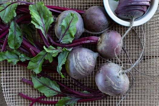 Bunch of fresh, spring, organic beetroot on kitchen grid, with slices of beetroot in ceramic plate on old wooden background. Top view. Rustic, dramatic, organic kitchen. Ingredients, menu. Healthy life concept