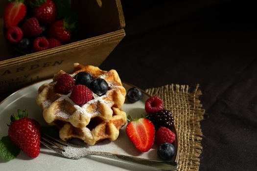 Belgian waffles with strawberries, summer berries on plate with canvas on dark old wooden dramatic background. Horizontal view. Menu, banner, poster, mock up. Rustic vintage style of kitchen.