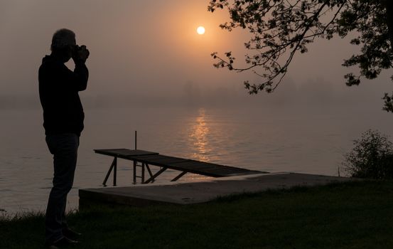 silhouette photographer at wooden jetty during sunset in the early morning over the river maas in limburg in holland with the trees mist and hazy fog