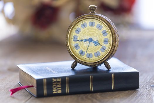 Old alarm clock on Holy bible with flowers on wooden table background